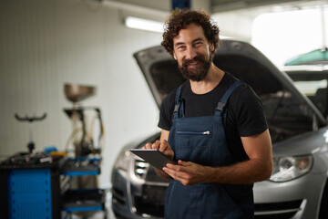 A skilled car mechanic stands in a garage, smiling while holding a tablet. The engine of a vehicle is open in the background, indicating ongoing repairs.