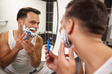 In a bright bathroom, a man is carefully shaving his face with a blue razor. He smiles at his reflection, enjoying his morning routine while applying shaving cream and preparing for the day.
