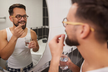 A man stands in front of a mirror in his bathroom, smiling as he prepares to take his medicine with a glass of water in the morning light.