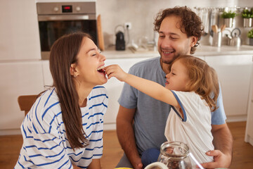 A family is gathered in the kitchen, sharing laughter and love as a child playfully feeds a piece of food to a smiling parent. The atmosphere is warm and cheerful.