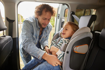 A father joyfully assists his child in securing the seatbelt in a car seat, ensuring safety before heading out for a family adventure on a sunny day.