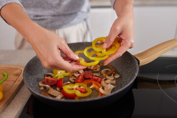 In a bright kitchen, a woman skillfully adds yellow bell pepper slices to a hot pan filled with mushrooms and red tomatoes, showcasing a vibrant cooking scene.