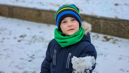 A little boy in a bright hat and green scarf plays with snow outside.
