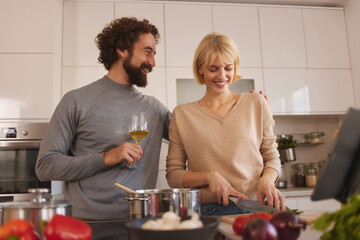 In a bright kitchen, a man smiles at a woman as she chops vegetables. They are enjoying wine while cooking, creating a warm and relaxed atmosphere filled with laughter and teamwork.
