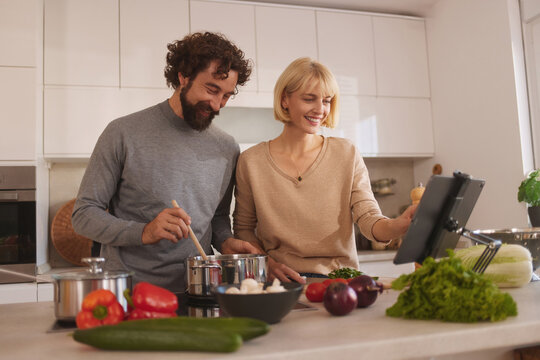 In a bright and airy kitchen, a couple joyfully cooks together. They are focused on a tablet displaying a recipe, surrounded by fresh vegetables and cooking utensils, creating an enjoyable atmosphere.