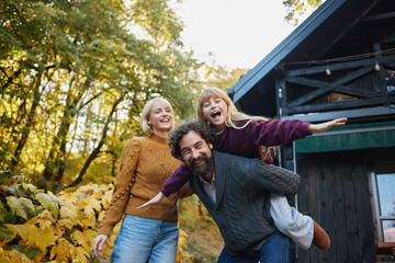 A joyful family stands outside a cozy cabin, with the mother and daughter laughing while the father gives his daughter a piggyback ride. Autumn leaves create a colorful backdrop.