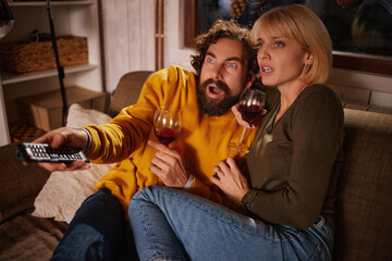 A couple sits closely on a sofa, engrossed in a tense movie scene. They hold glasses of wine, showing excitement and surprise as they watch the screen. © Stockphotodirectors