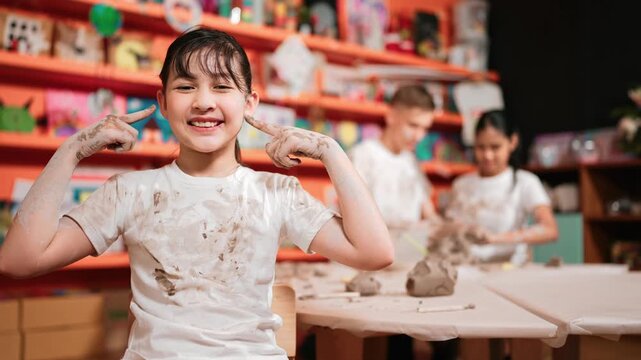 Asian girl pose at camera while diverse children modeling clay behind. Happy cute student wearing dirty shirt while looking at camera at workshop in art lesson. Blurring background. Edification