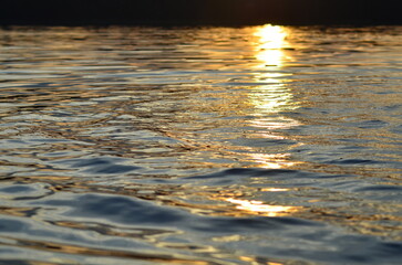 A large lake at sunset. A sunny evening. The sky is covered with clouds, through which the sun shines. A wide lake with slight ripples on the surface reflects the sun's rays, giving water a golden hue