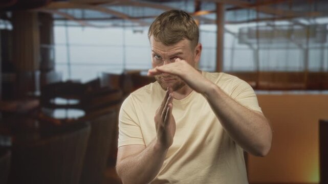 Man making timeout gesture with hands in hotel lounge, wearing beige tshirt and beard, looking serious; boundary warning.