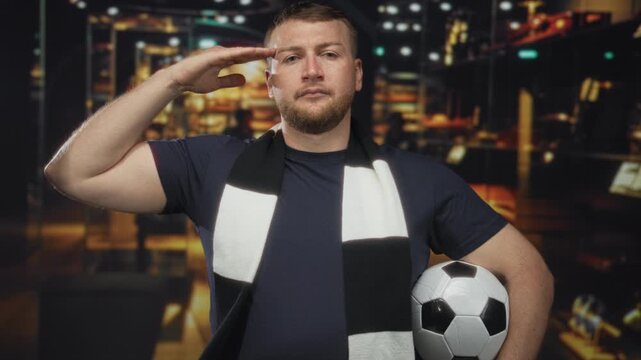 Man salutes hand to forehead holding a soccer ball in a shop building with black and white scarf; pride fandom loyalty.