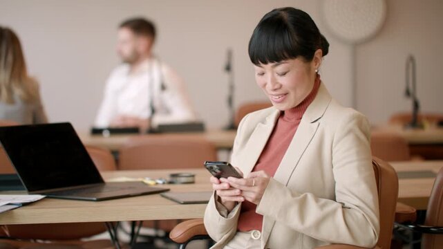 Japanese woman in a stylish office shares a joyful moment while checking her phone. Her smile brightens the workspace as colleagues focus on tasks.