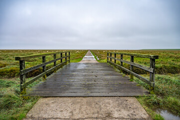 A wooden bridge stretches into a vast, grassy field under a cloudy sky, leading to a distant path, North Sea, St Peter Ording, Germany