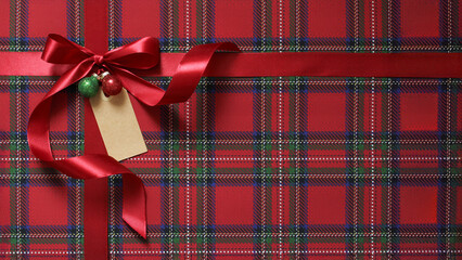 Christmas present, gift close-up. Red  satin ribbon, bow with green and red ornament and empty tag on plaid paper background.