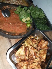 Fried chicken pieces, traditional Indonesian sambal in a stone mortar, fresh lime, basil leaves, long beans, and other vegetables arranged on a wooden table. Suitable for themes of Indonesian cuisine.