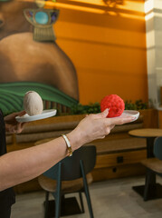 Close-up image of two hands holding white plates with creative gourmet desserts &mdash; one red raspberry shaped dessert and one pistachio inspired dessert.