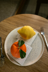 Top view of two creative gourmet desserts shaped like a lemon and an orange, placed on a ceramic plate on a wooden table.