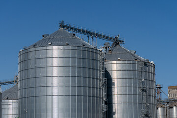 Steel grain silos stand tall against a clear blue sky in an agricultural setting during daylight hours