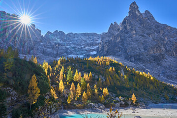 Mount Sorapiss towers above Lake Sorapis and forms the dramatic backdrop visible from lake&rsquo;s shore. Turquoise color of the lake. Autumn landscape with golden larch trees. Rays radiating from the sun.