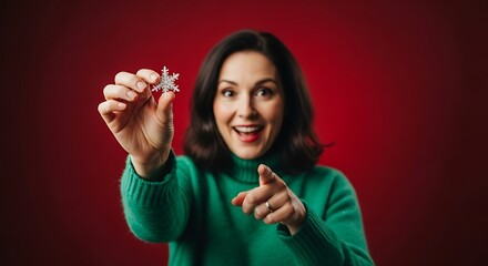Woman in green sweater holding snowflake against a red background while pointing at the camera