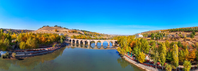 Panoramic view of the historic On Gözlü Köprü (Ten Arches Bridge) near Diyarbakır, Turkey, spanning the Tigris River, surrounded by trees with autumn foliage under a bright blue sky 