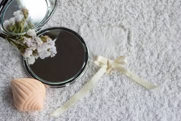 Compact mirror reflecting delicate flowers lying on a plush white towel with a decorative soap bar and a cream ribbon, symbolizing beauty, self care, and a calming spa atmosphere