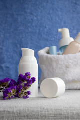 Skincare products arranged on a folded towel, pump lotion, white cosmetic jar and bottles with purple dried flowers on gray linen against a textured blue backdrop, serene spa vibe