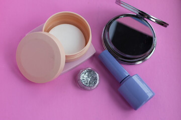 Cosmetics arrangement on a vibrant pink background featuring a compact mirror, loose glitter, a container of nail polish remover wipes, and nail polish, symbolizing beauty and self care