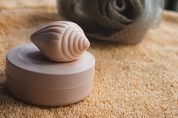 Seashell shaped soap sitting on a pink cream container on a soft towel, creating a serene and inviting atmosphere for personal beauty, hygiene, and wellness rituals