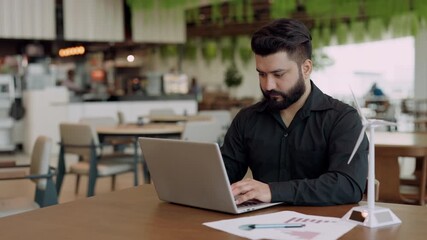 Portrait of young Indian engineer with miniature windmill types on laptop in office. Power network expert promotes green energy generation technology in public coworking premise - Powered by Adobe