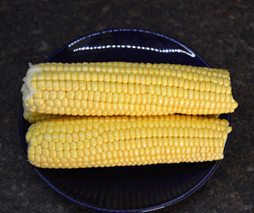 Raw corn on the cob on a dark plate. A delicious, ready-to-eat vegetable. A tasty and healthy dessert for both children and adults. A source of vitamins and fiber.