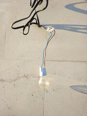 The light bulb hangs against a light concrete wall, illuminated by sunlight. Subtle shadows and traces of time are visible on the surface, creating an atmosphere of simplicity and everyday life.