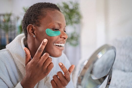 Young woman applying a hydrogel eye patch mask smiling at the mirror
