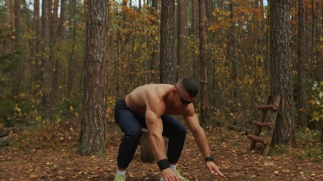 Young muscular man without shirt and wearing sunglasses performs burpee exercise in glade in an autumn forest. Tracking shooting. Calisthenics. Bodyweight exercises