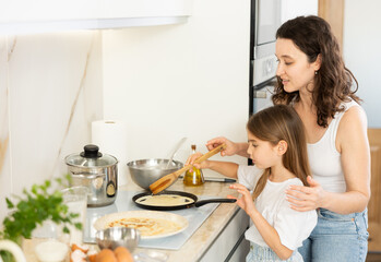 Happy mother and her preteen daughter frying pancakes on stove in the kitchen