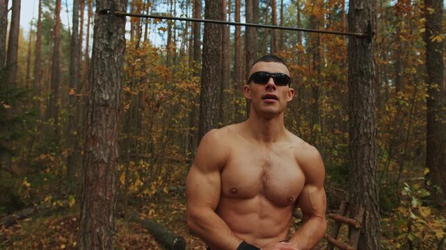 Shirtless young man stands in an autumn forest near horizontal bar between trees and showing off his powerful biceps and abs. Bodybuilding concept
