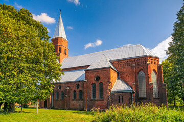 Historic Church of the Assumption of the Blessed Virgin Mary in Otynia, Ukraine. Red brick architecture with a silver spire, surrounded by lush green trees under a blue sky