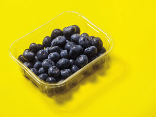 A plastic tray of blueberries sits on a bold yellow color background. The tray is clear and filled with fresh organic fruits ready for consumption. Healthy diet concept. Natural product.
