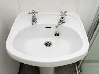 A white British style sink with two separate faucets and a drain. The sink is in a bathroom.