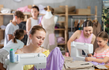 Enthusiastic teenage girl holding sewing work with smile during practical clothing construction course for school students led by experienced sewing instructor in training workshop....