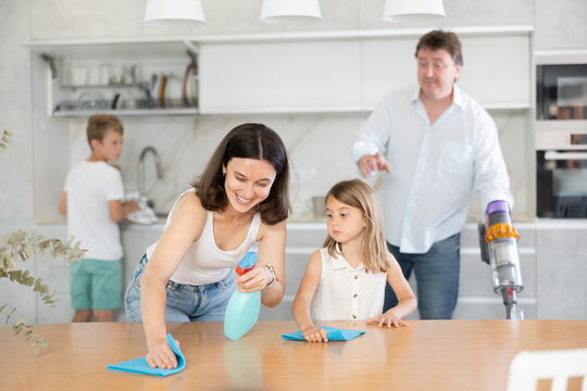 Little girl helps mother wash kitchen table. Husband and son are cleaning kitchen, washing dishes, vacuuming floor in background. Joint family house cleaning, household tasks, household help
