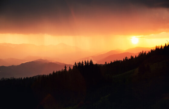 Sun sets behind distant mountains, casting warm hues across the sky during a heavy downpour illuminated by the sun's rays.
