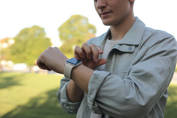 A young man looking down at his sleek smartwatch in a sunny, pleasant park setting, emphasizing time management, digital communication, and tracking personal health and fitness goals.