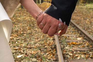 Newlyweds holding hands walking along old railroad tracks covered with autumn leaves. Close-up of groom and bride arms. Romantic wedding love and life journey concept