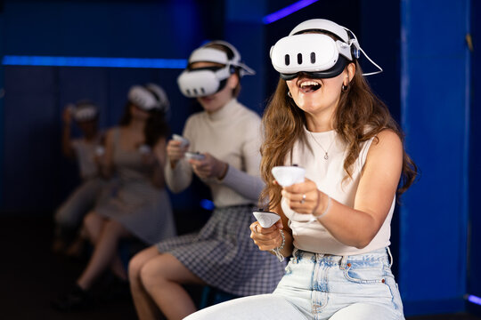 Cheerful emotional young female in VR goggles sitting in virtual reality room with group of friends, manipulating objects or shooting at targets with gaming controllers in hands..