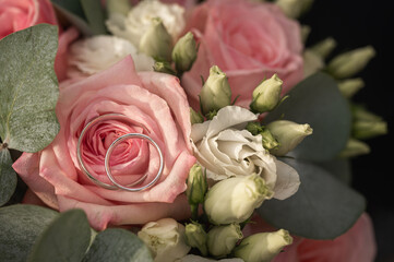 Close-up of two silver wedding rings resting on pink rose petals in a bridal bouquet with white flowers and eucalyptus leaves. Romantic symbol of love and marriage, selective focus