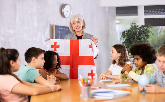 Woman high school teacher, conducting a lesson in the classroom, tells pupils the history of Georgia and holds the national .flag of the country