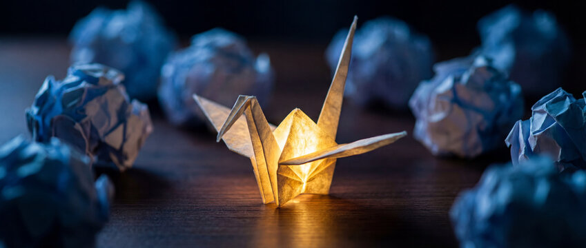 Glowing origami crane standing out among crumpled blue paper balls on a dark wooden desk, symbolizing innovation and the creative process. - Powered by Adobe