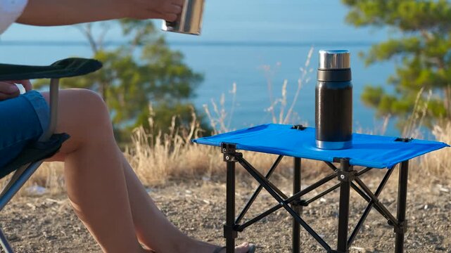 A woman drinks a hot drink by the sea. Tourist sits near the sea, pouring a hot beverage from a thermos into a metal mug. A blue folding table and chair stand against the backdrop of a calm sea