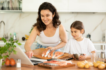 Mom and daughter are preparing festive salmon dish for family dinner. While cooking, mom peeks at laptop screen, clarifies recipe.